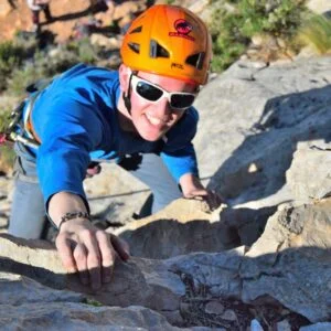 Rock climber wearing helmet, sunglasses, and harness climbing in the sun.
