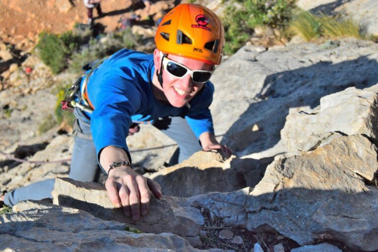 Rock climber wearing helmet, sunglasses, and harness climbing in the sun.