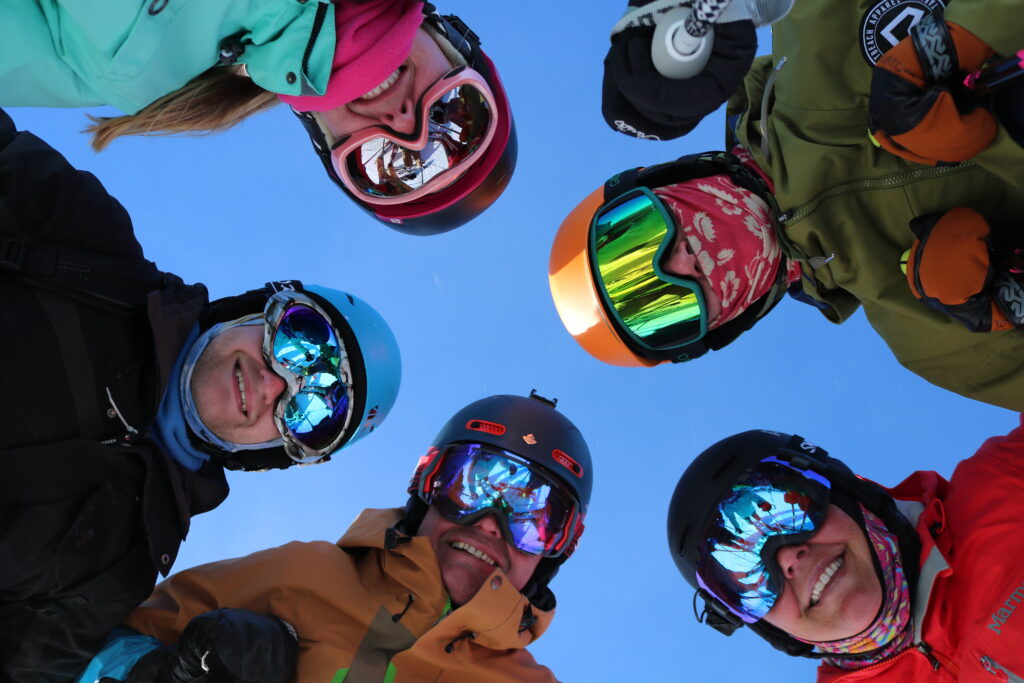 People smiling wearing ski gear, looking down at the camera with blue sky above.